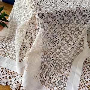 Op de afbeelding: Een wit kanten tafelkleed met een herhalend cirkelvormig geometrisch patroon in het midden. De stof bevat een effen witte rand en een decoratieve geschulpte afwerking met een bloemmotief langs de omtrek van het textiel.