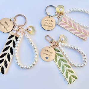 Peut inclure: Porte-clés dorés avec des lanières décoratives et des perles. Un porte-clés a une lanière avec une broderie de feuilles noires et le texte "Merci maîtresse". Un autre a une lanière avec une broderie de feuilles roses et le texte "Veux-tu être ma Marraine ?". Le troisième a une lanière avec une broderie de feuilles vertes et le texte "Maman d'amour".
