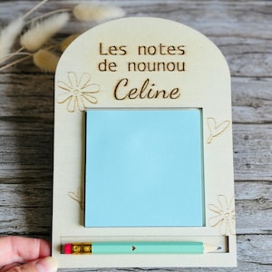 Peut inclure: Tableau mémo en bois avec le texte "Les notes de nounou Celine". Il comprend un post-it bleu clair et un crayon vert clair. Des motifs floraux et en forme de cœur sont gravés dans le bois.