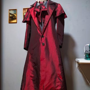 May include: A long, burgundy coat with a high collar and two black buttons. The coat has a shiny, textured appearance and is paired with a white collared shirt and a bow tie. The outfit is displayed on a mannequin.