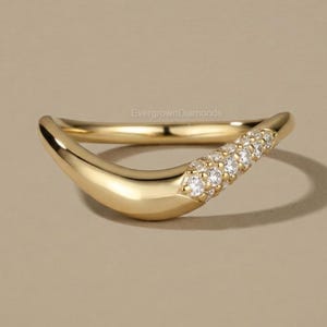 Puede incluir: Un anillo de oro curvado con una fila de pequeñas piedras preciosas claras engastadas en la parte superior. El anillo tiene un acabado liso y pulido y un diseño moderno. El anillo está sobre un fondo neutro.