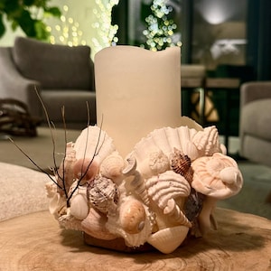 Könnte beinhalten: Eine weiße Kerze steht in einem Halter aus Muscheln. Der Halter besteht aus einer Vielzahl von Muscheln, darunter große Jakobsmuscheln, kleinere Conch-Muscheln und mehrere kleinere, zartere Muscheln. Der Kerzenhalter steht auf einer Holzoberfläche.