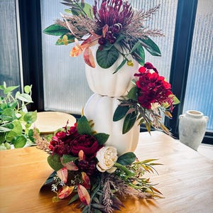 May include: A stacked decorative arrangement featuring two matte white pumpkins adorned with deep red and burgundy floral accents, including roses, hydrangeas, and dark foliage, creating a layered centrepiece with varied textures and autumn-toned leaves.