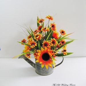 May include: A rustic galvanized metal watering can filled with a bouquet of faux sunflowers and daisies in shades of orange, yellow, and red. The flowers are arranged in a cascading style, creating a charming and colorful centerpiece.