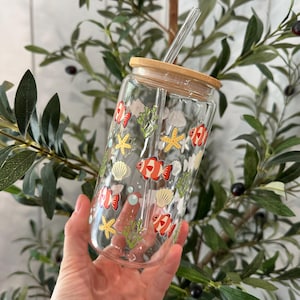 May include: A clear glass reusable water bottle with a bamboo lid and a straw. The bottle has a colorful design of orange and white fish, seashells, and seaweed.