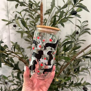 May include: A clear glass tumbler with a bamboo lid and a clear straw. The tumbler is decorated with black dachshund silhouettes and red heart shapes. The tumbler is held in front of a background of green leaves and a white wall.