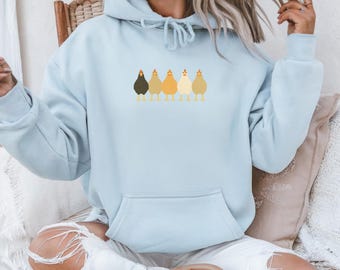 Puede incluir: Sudadera con capucha azul claro con bolsillo delantero y capucha con cordón. La sudadera presenta una fila de seis pollos de dibujos animados en varios tonos de marrón y amarillo en el pecho. Los pollos están uno al lado del otro.