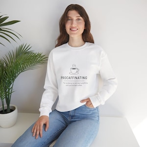 Puede incluir: Sudadera blanca de manga larga con la palabra "PROCAFFINATING" y un dibujo de una taza de café. El texto de abajo dice: "La tendencia a no empezar nada hasta que hayas tomado café."