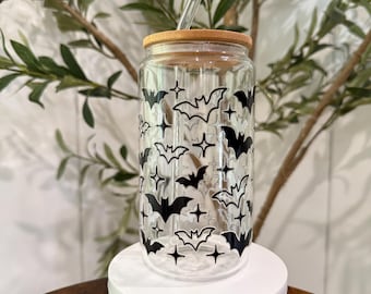 May include: A clear glass tumbler with a bamboo lid and a clear straw. The tumbler is decorated with a pattern of black bats and stars. The tumbler is sitting on a white pedestal.