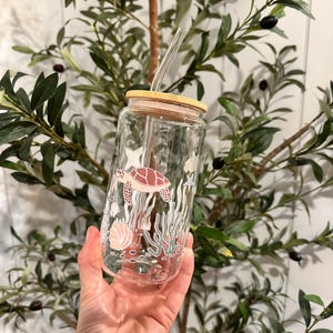 Può includere: Un bicchiere di vetro trasparente con coperchio in bambù e cannuccia trasparente. Il bicchiere presenta un design a tema marino con una tartaruga marina rosa, coralli e altre creature marine. Il bicchiere è tenuto davanti a uno sfondo verde foglioso.