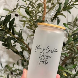 May include: Frosted glass can-shaped tumbler with a bamboo lid and a clear glass straw. The tumbler has the text "Your Custom Design Here" in a script font. The background features green foliage.