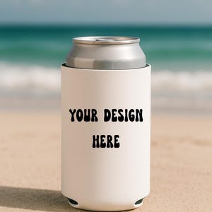 May include: A white can cooler with the text "YOUR DESIGN HERE" in black, holding a silver can. The cooler is on a sandy beach with the ocean in the background.