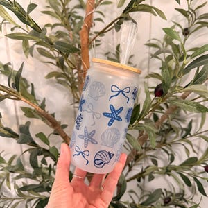 May include: A frosted glass can-shaped tumbler with a bamboo lid and straw. The tumbler is decorated with blue seashell, starfish, and bow designs. The background features olive branches.