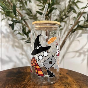 May include: A clear glass reusable water bottle with a bamboo lid and a straw. The bottle has a variety of stickers on it, including a witch's hat, glasses, a scarf, a feather, and a potion bottle.