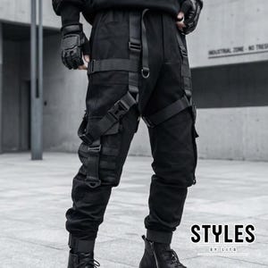 Op de afbeelding: Zwarte cargobroek met meerdere zakken en riemen. Het tactische ontwerp omvat verstelbare riemen en gespen. De tekst "STYLES by LITA" staat in het wit afgebeeld.