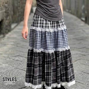 May include: A long, tiered skirt featuring alternating panels of black and white plaid and gingham patterns. The skirt is accented with horizontal bands of white lace trim between each tier and along the bottom hem, paired with a dark grey top.