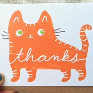 May include: A greeting card featuring an orange cat illustration with the word "thanks" in white script. The cat has green eyes and black whiskers. The card is on a brown envelope.