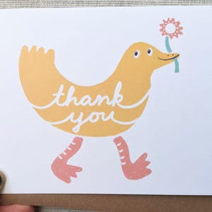 May include: A white greeting card with a cartoon duck illustration. The duck is yellow with pink feet and is holding a flower. The words "thank you" are written in cursive on the duck. The card is on a brown envelope.