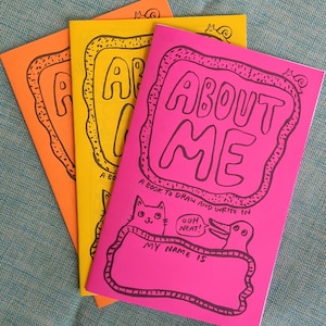 May include: Three pink, yellow, and orange paperbacks with the title "About Me" and a black and white illustration of a cat and a duck. The book is a "book to draw and write in".