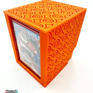 May include: An orange, cube-shaped card holder with a decorative swirl pattern. The holder is open on one side, revealing a card with a colorful illustration. The Print and Chill 3D logo is visible in the bottom left corner.