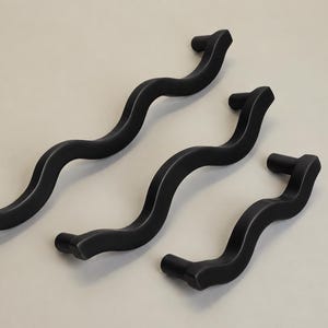 May include: Three matte black cabinet pulls arranged diagonally, each featuring a distinct wavy, serpentine shape with two mounting points at the ends. The handles vary in length, showcasing a consistent dark finish and a smooth, curved design.