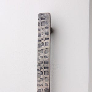 May include: A rectangular, textured metal door pull with a dark grey finish. The surface has a pattern of recessed squares. The door pull is mounted on a white wall, providing a minimalist aesthetic.
