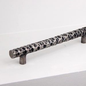May include: A hammered metal cabinet pull with a textured surface. The handle is a dark gray color with a cylindrical shape and two short posts for mounting. The hardware is designed for use on drawers or cabinet doors.