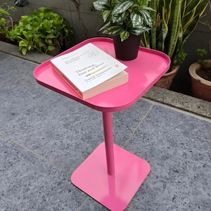 May include: A vibrant pink side table with a square top and base, joined by a thin, cylindrical stem. A potted plant and a book titled "Thinking, Fast and Slow" are on the table. The table is about 46 cm high.