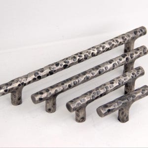 Könnte beinhalten: Satz von vier gehämmerten Metall-Schrankgriffen in verschiedenen Größen. Die Griffe haben ein strukturiertes, gesprenkeltes Aussehen mit dunkler grauer Oberfläche. Die Griffe sind T-förmig, mit einem Stab und zwei kurzen Beinen.