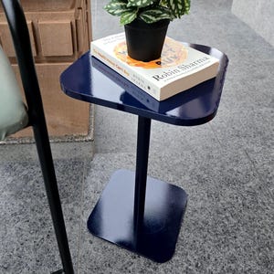 May include: A navy blue side table with a square base and top, supporting a small potted plant and a book titled "The Wealth Money Can't Buy" by Robin Sharma. The table is made of metal and has a glossy finish.