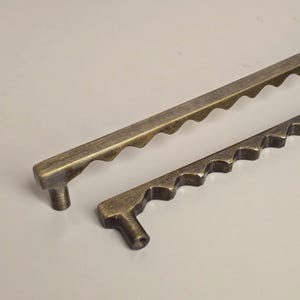 May include: Two antique bronze-colored cabinet pulls. The pulls have a unique design with a series of wave-like cutouts along the top edge. One pull is approximately 10 inches long, and the other is about 6 inches long.