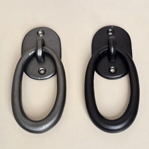 Op de afbeelding: Twee metalen ovale handgrepen. Eén is donkergrijs, de andere zwart. Elke handgreep heeft een ovale ring bevestigd aan een ovale achterplaat met twee schroefgaten voor montage. De achtergrond is lichtbeige.