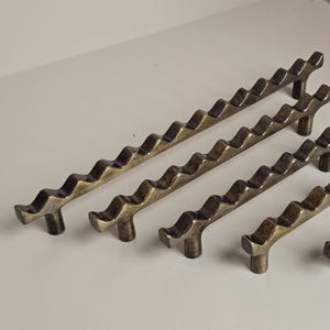 Puede incluir: Un conjunto de tiradores y pomos de armario de color bronce. Los tiradores tienen un diseño ondulado, y los pomos son versiones más pequeñas de los tiradores. El hardware está dispuesto sobre una superficie blanca.