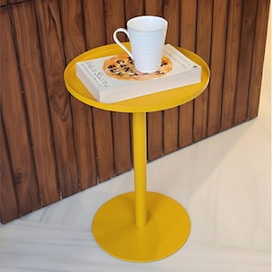 Peut inclure: Une table d'appoint jaune vif avec un plateau et une base ronds. Une tasse blanche et un livre intitulé "CAN'T BUY" sont sur la table. La table a un seul pied droit. Le fond présente un mur en bois.