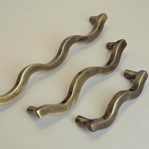 Puede incluir: Tres tiradores de armario de latón antiguo con un diseño ondulado y orgánico. Los tiradores tienen un acabado cálido y envejecido y están dispuestos sobre un fondo claro. El tirador más grande mide unos 20 cm de largo.