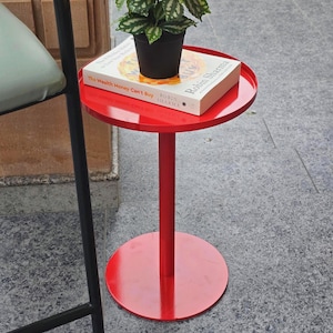 Może przedstawiać: Mały, okrągły, czerwony stolik boczny z centralną nogą i okrągłą podstawą. Na stole stoi roślina doniczkowa z zielono-białymi liśćmi oraz książka zatytułowana "The Wealth Money Can't Buy" autorstwa Robina Sharma.