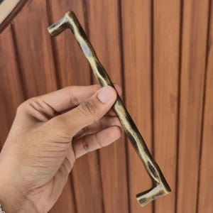 May include: A bronze-colored cabinet pull with a rustic, branch-like design. The handle is held against a brown wooden background. The pull is approximately 10 inches long.