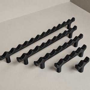 May include: A collection of five matte black cabinet pulls arranged by size from longest to shortest. Each pull features a distinct wavy, undulating top edge and is supported by two cylindrical posts, except for the smallest single-post pull.