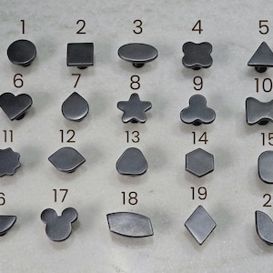 Op de afbeelding: Een verzameling van twintig donkergrijze kastknoppen in verschillende vormen, waaronder cirkels, vierkanten, harten, sterren en geometrische ontwerpen. Elke knop is genummerd van 1 tot 20, wat een reeks decoratieve opties biedt voor meubels of kasten.