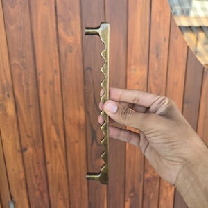 May include: A bronze-colored door pull with a unique, jagged edge design. The handle is held against a wooden door, showcasing its decorative and functional aspects. The pull has a length of approximately 12 inches.