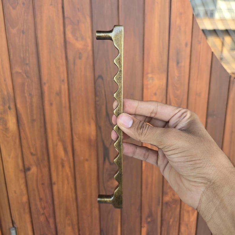 May include: A bronze-colored door pull with a unique, jagged edge design. The handle is held against a wooden door, showcasing its decorative and functional aspects. The pull has a length of approximately 12 inches.