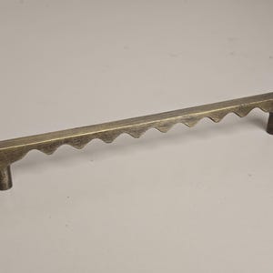 May include: A bronze-colored cabinet pull with a unique design. The handle is rectangular with a series of inverted, rounded triangles along the top edge. It has two cylindrical posts for mounting.