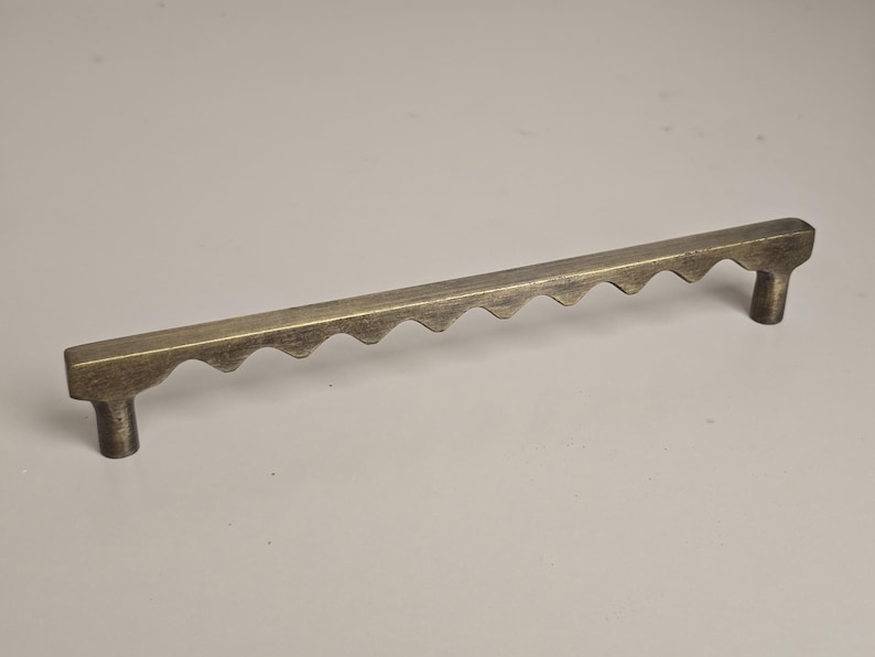 May include: A bronze-colored cabinet pull with a unique design. The handle is rectangular with a series of inverted, rounded triangles along the top edge. It has two cylindrical posts for mounting.