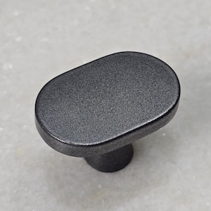 Può includere: Un pomello per armadio grigio scuro, di forma ovale, con una superficie strutturata. Il pomello ha una parte superiore arrotondata e una base cilindrica corta. L'hardware è in metallo ed è adatto per cassetti o armadi.