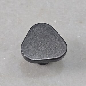 Könnte beinhalten: Dunkelgrauer, dreieckiger Schrankknopf mit strukturierter Oberfläche. Der Knopf hat eine abgerundete, ergonomische Form und einen kleinen, kreisförmigen Sockel. Geeignet für Schubladen, Schränke und Möbel.