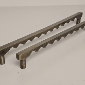 May include: Two antique bronze cabinet pulls. The pulls have a rectangular shape with a series of wave-like cutouts along the top edge. Each pull has a cylindrical post on each end for mounting. The pulls are different sizes.