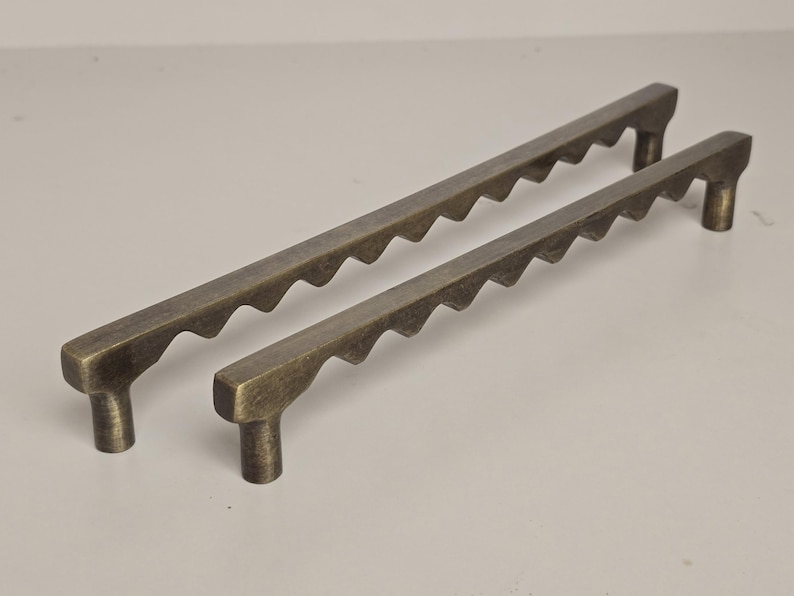 May include: Two antique bronze cabinet pulls. The pulls have a rectangular shape with a series of wave-like cutouts along the top edge. Each pull has a cylindrical post on each end for mounting. The pulls are different sizes.