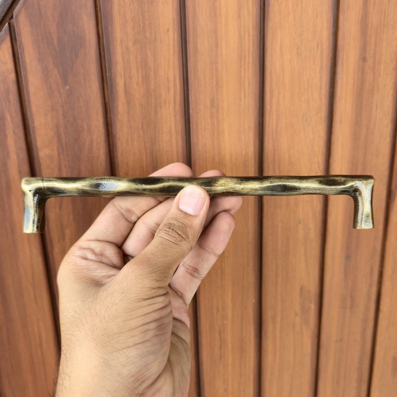 May include: A bronze-colored cabinet pull with a textured, organic design. The handle is held against a wooden door. The pull has a rectangular shape with a curved, textured bar and right-angled ends. The hardware is approximately 8 inches long.