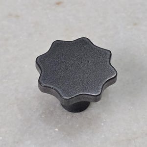 Könnte beinhalten: Ein dunkelgrauer, sternförmiger Knopf mit strukturierter Oberfläche. Der Knopf hat einen runden Sockel und wird wahrscheinlich für Möbel oder Maschinen verwendet. Das Objekt befindet sich vor einem hellgrauen Hintergrund.