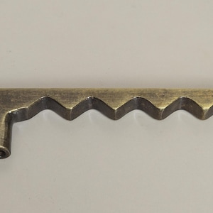 May include: A bronze-colored cabinet pull with a unique, wavy design. The hardware features a series of zig-zag patterns along its length, with a small, cylindrical projection on one end for mounting. This decorative pull adds a vintage touch to furniture.
